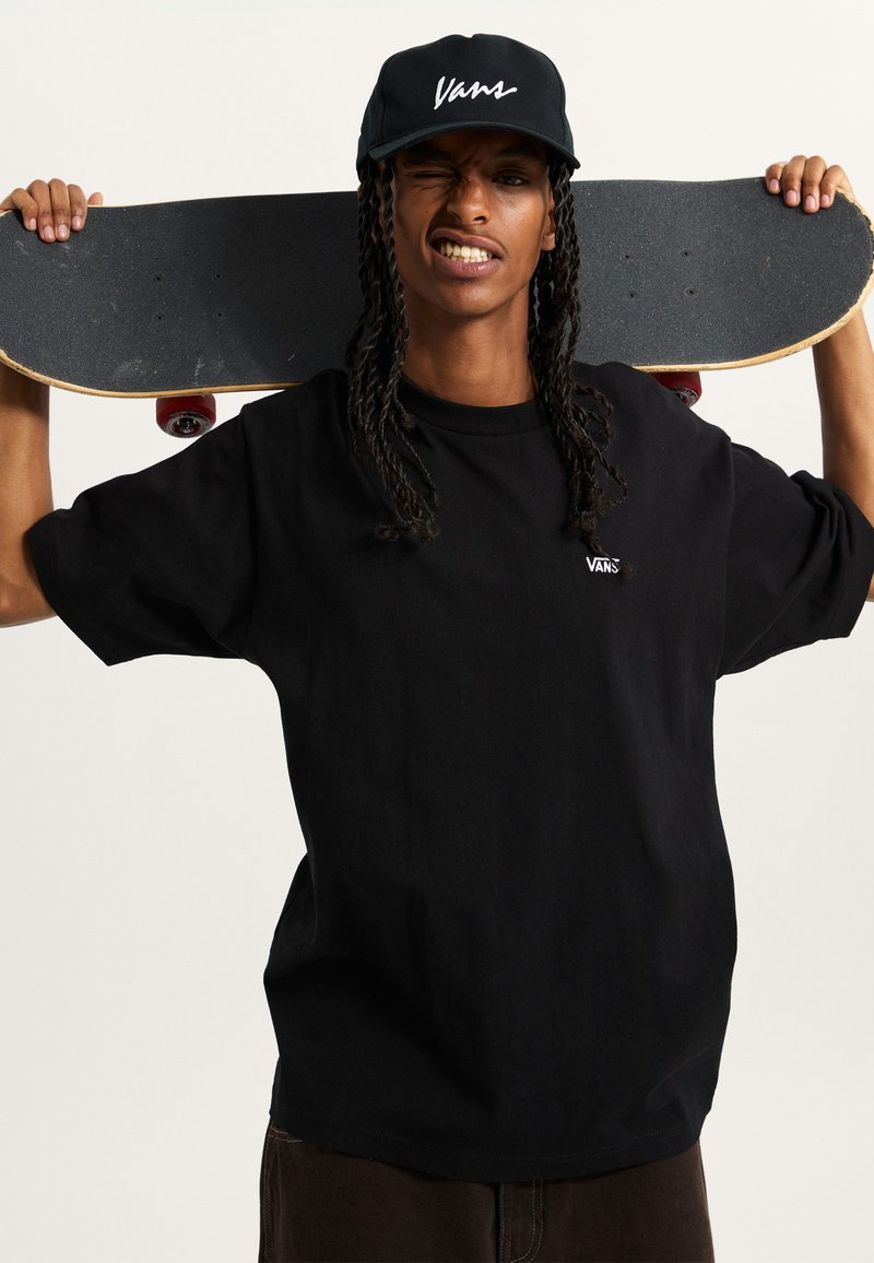 Schwarzes T-Shirt mit gesticktem "Vans"-Logo, kombiniert mit einer schwarzen Mütze und einem Skateboard, das über die Schulter gehalten wird und eine strukturierte Grifffläche aufweist.