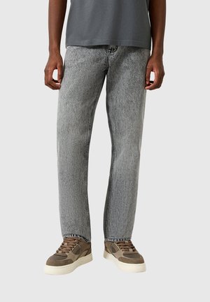 Partie inférieure du corps d'une personne portant un jean droit gris clair, des baskets marron et beige, et un t-shirt gris foncé sur un fond uni.