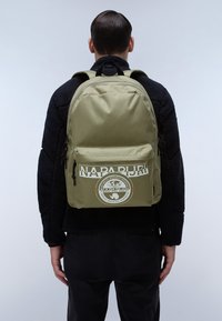 Zaino verde oliva con una tasca frontale, caratterizzato da un logo e testo bianchi. Realizzato in materiale resistente, ha spallacci regolabili e una maniglia superiore.