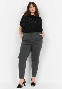 T-shirt noir avec un motif argenté ondulé, pantalon gris ajusté et sandales à talons noires avec des accents floraux. Tissu lisse et silhouette ajustée.