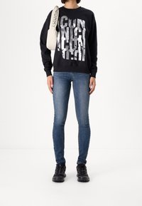 Sudadera negra con un gran texto gráfico plateado, combinada con jeans ajustados azules y zapatos negros con estampado. Bolso crema claro con logo.