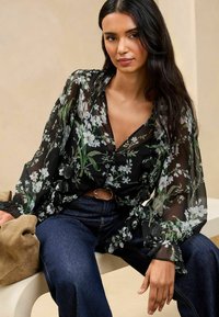 Schwarze transparente Bluse mit floralen Mustern in Weiß und Grün, ausgestattet mit langen Ärmeln und gerafften Manschetten, kombiniert mit dunklen Jeans.