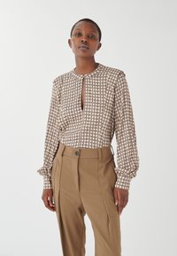 Blusa marrone a fantasia con scollo a chiave, abbinata a pantaloni beigi alti in vita con chiusura a bottone e tasche laterali.