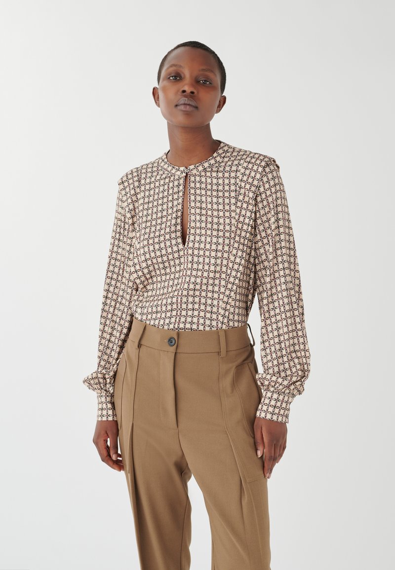 Blusa marrone a fantasia con scollo a chiave, abbinata a pantaloni beigi alti in vita con chiusura a bottone e tasche laterali.