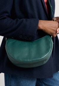 Borsa a tracolla verde in pelle, dalla forma arrotondata, texture liscia e hardware minimale, appoggiata su un cappotto blu navy e jeans di denim.