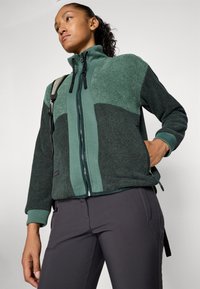 Woman wearing a green and dark gray zip-up fleece jacket with hand in pocket, carrying a backpack strap over one shoulder against a plain background.