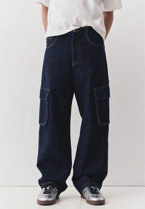 Person trägt dunkelblaue, weit geschnittene Cargo-Jeans und silberne Turnschuhe, steht vor einer hellen einfarbigen Wand.