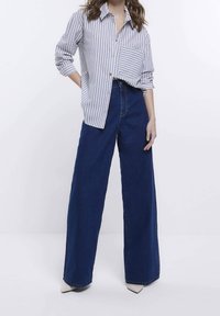 Pasiasta koszula z guzikami w jasnoniebieskim i białym kolorze, zestawiona z wysokim stanem szerokimi dżinsami w ciemnym odcieniu i białymi szpilkami o spiczastym nosku.