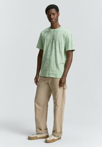 Ljusgrön t-shirt med strukturerat mönster, rund halsringning, kort ärm, i kombination med beige cargobyxor och off-white sneakers.