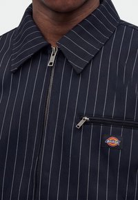 Κοντινό πλάνο ατόμου που φοράει σκούρο μπλε σακάκι με ρίγες, ασημένιο φερμουάρ και τσέπη στο στήθος με φερμουάρ με το σήμα της Dickies.