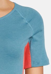 Top sportivo blu a maniche corte, con un pannello laterale rosso e tessuto testurizzato, scollo rotondo e dettagli di cucitura visibili.