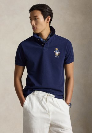 Mand iført navy polotrøje med broderet bjørn og hvide bukser, stående med hænderne i lommerne og kiggende til siden.