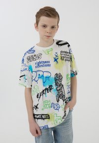 Maglietta bianca con una combinazione di grafiche colorate in stile graffiti nei colori blu, verde e nero, abbinata a jeans di denim chiaro.