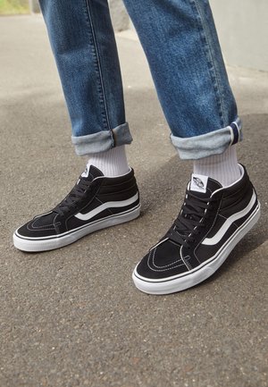 Personne portant des baskets montantes noires et blanches, des chaussettes blanches et un jean bleu retroussé, debout sur un revêtement en asphalte.