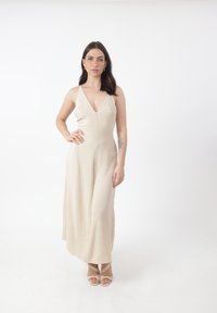 Beige maxi kjole uden ærmer med V-hals og blød, tekstureret stof. Sammen med lyse beige stroppsandaler. Simpelt og elegant design.