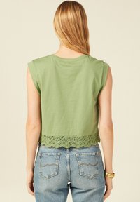Top verde in cotone cropped con maniche a cappuccio, caratterizzato da un orlo in pizzo scallop. Abbinato a jeans blu a vita alta.