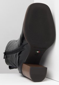 Botte en cuir verni noir avec fermeture éclair, semelle en caoutchouc texturée et talon en bois. Nom de la marque et pays d'origine sur la semelle.