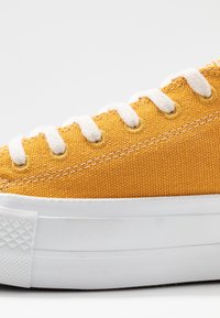 Keltainen kangaskengä, jossa on valkoiset nauhat, teksturoitu päällinen ja valkoinen kumipohja, jossa on timanttimuotoilu kärjessä.