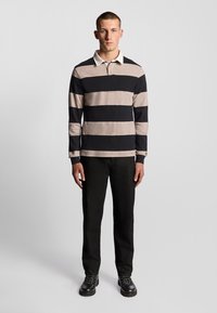 Langarm-Polohemd in beige und schwarzen Streifen, mit einem weißen Kragen. Kombiniert mit schwarzen Hosen und schwarzen Schnürstiefeln.