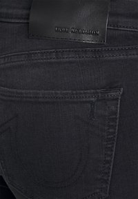Vaqueros de mezclilla negros con una textura suave, con una etiqueta de cuero etiquetada como "TRUE RELIGION", y un bolsillo trasero con costuras detalladas.