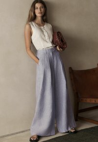 Lätt krämfärgad utan ärmar-top med knappdetaljer, i kombination med högmidjade vida lavendelfärgade byxor och en texturerad bordeauxfärgad clutch.