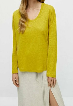 Woman wearing a mustard yellow long-sleeve top and a light beige skirt with a front slit, standing against a plain background.