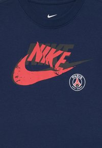 Námornícky modré bavlnené tričko s veľkým červeným logom Nike s čiernym obrysom a malým okrúhlym logom Paris Saint-Germain na spodnej strane.