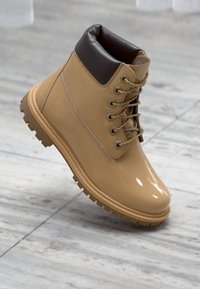 Stivale impermeabile beige con finitura lucida, colletto imbottito marrone, occhielli in metallo e suola rugosa per una migliore aderenza.