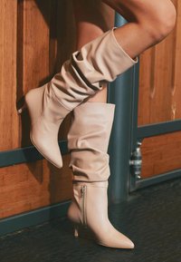Botine din piele bej, până la genunchi, cu un design lejer, fermoare laterale și tocuri tip kitten, purtate de o persoană care stă în picioare lângă un perete din lemn.
