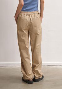 Beige relaxed-fit pants with an elastic waistband, two back pockets, and a smooth texture, paired with black slip-on shoes.