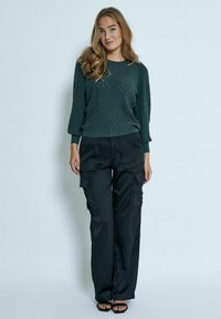 Maglione texturizzato verde scuro con orlo a coste, abbinato a pantaloni cargo neri dal fit morbido con tasche laterali. Il modello è in piedi su uno sfondo neutro.