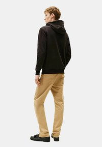 Sort hættebluse med ribbede manchetter, parret med lyse khaki bukser. Modellen har sorte sko på og ses bagfra mod en neutral baggrund.
