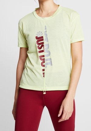 Femme portant un t-shirt jaune clair à manches courtes avec un texte vertical rouge et gris "JUST DO IT" et des leggings rouges, debout devant un fond uni.