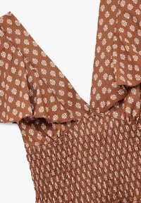 Brun mønstret kjole med hvid blomstermotiv, der har bølgede ærmer og smock-detalje ved kroppen, lavet af tekstureret stof.