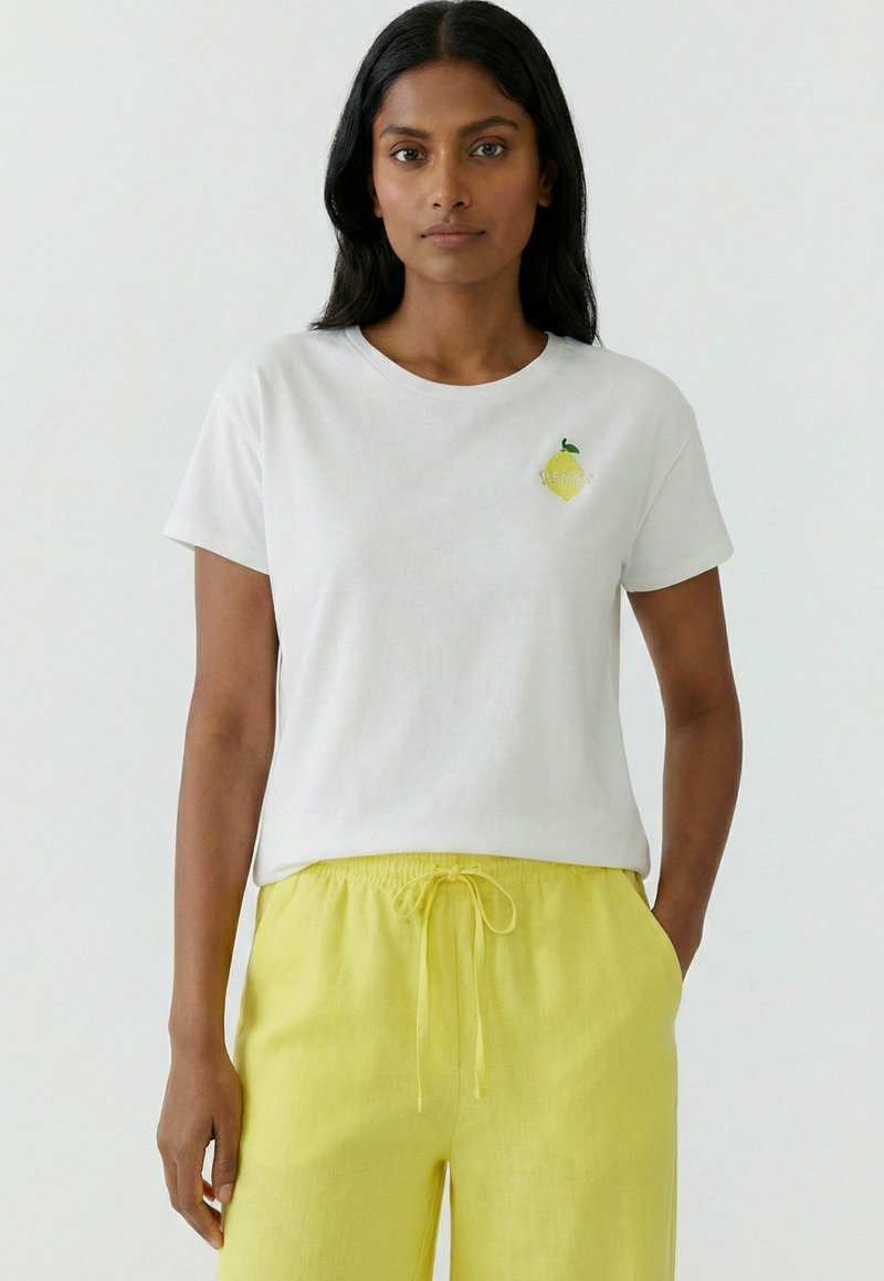 Woman wearing white t-shirt with small lemon graphic and yellow drawstring pants standing against plain white background.