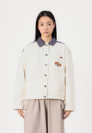 Veste couleur crème avec un col gris, comportant deux poches poitrine, des boutons argentés et un motif brodé sur la poche gauche.