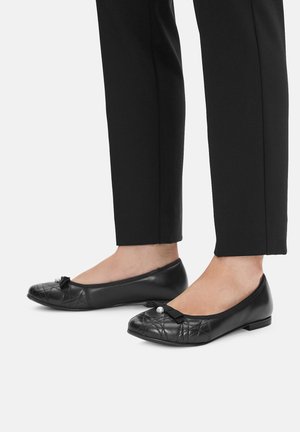 Czarne płaskie buty z pikowanym wzorem, małą kokardką i perłowym akcentem na palcu, zestawione z czarnymi spodniami.