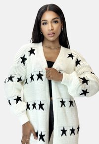 Cardigan lavorato a maglia in crema con motivi a stelle nere. Presenta maniche lunghe, apertura frontale e una vestibilità rilassata. Dettagli in tessuto testurizzato evidenti.