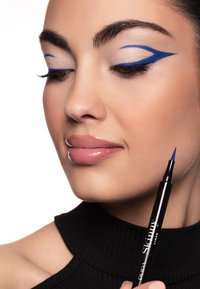Penna per eyeliner blu tenuta vicino al volto di una modella; caratteristiche includono un audace eyeliner alato blu, pelle liscia e labbra nude lucide. Top nero con trama.