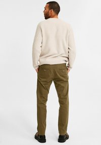 Beige gebreide trui met gestructureerde mouwen, gecombineerd met olijfgroene broeken met dubbele achterzakken. Het model draagt zwarte schoenen.