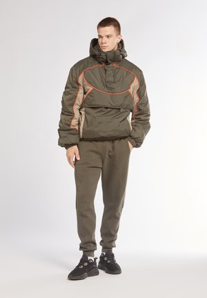 Giacca a vento verde oliva con cuciture arancioni, colletto alto e zip frontale. Abbinata a pantaloni jogger verdi oliva e sneakers nere.