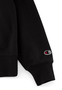 Sudadera negra con puños y dobladillo acanalados, hecha de material de felpa; incluye un pequeño parche de logo en rojo y blanco en la manga.