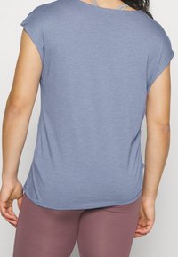 Person som bär en lös blå kortärmad topp över en grå sportbehå och tajta mauvefärgade leggings, sedd bakifrån.