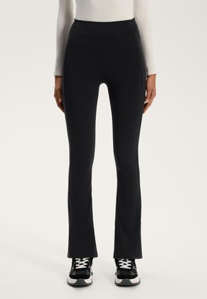 High-waisted black leggings with a stretchy fit, flared legs, seam details, and two side pockets; paired with black and white sneakers.