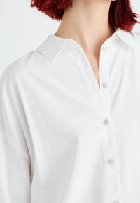 Personne aux cheveux roux portant une chemise blanche boutonnée avec un col visible, cadrée au niveau du cou et des épaules sur fond blanc.