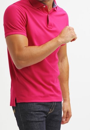 Mann trägt hellpinkes kurzärmeliges Poloshirt und dunkle Jeans, mit einem angewinkelten Arm und Faust nahe der Brust vor weißem Hintergrund.