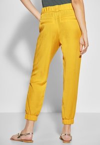 Gele, lichte broek met een elastische tailleband en enkelboorden. Heeft een soepele structuur en twee achterzakken.