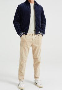 Marinblå corduroy bomberjacka med randiga ärmslut, beige corduroybyxor och vita sneakers med svarta detaljer. Avslappnad, lös passform.