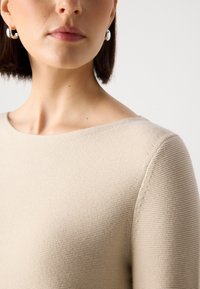 Suéter de tricô bege com um decote largo, tecido texturizado e acabamento suave. Brincos de argola prateados complementam o conjunto.