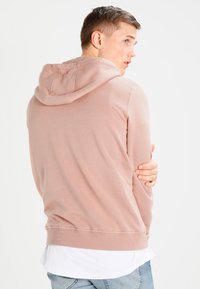 Lys rosa hettegenser laget av myk bomull, med snor i hetten, ribbestrikkede mansjetter og kant; brukt over en hvit skjorte; bakfra.